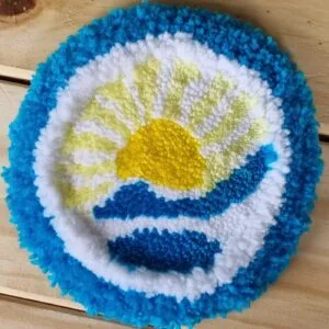 Alfombrita posavasos Argentina hecha a mano, con diseño circular de sol y montañas en celeste y blanco inspirado en símbolos Patrios.